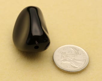 May include: A smooth, polished, black, teardrop-shaped stone bead with a single drilled hole at the base. The bead is positioned next to a Canadian 25-cent coin for scale, which has a diameter of approximately 0.94 inches, to show its size.