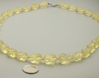 May include: A necklace composed of translucent, pale yellow, faceted beads with a twisted, geometric shape. The beads are strung together with small, light-colored spacers. A silver-toned metal clasp secures the ends. A US quarter coin is placed nearby for scale.