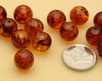 May include: A collection of round, translucent amber-colored beads with swirling dark brown inclusions. Each bead features a small center hole for stringing. A silver-colored Canadian 25-cent coin is placed among the beads for scale reference.
