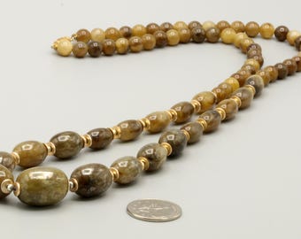 May include: A necklace featuring a strand of polished, variegated brown and tan oval beads separated by small gold-toned metal spacers. The beads vary in size, with larger beads at the front and smaller beads toward the back, shown next to a US quarter coin.