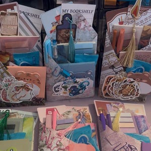 May include: Assortment of decorative paper organizers filled with stationery, stickers, and pens. Each organizer features a unique design, including animal illustrations, tassels, and the text "MY BOOKSHELF". The color palette includes pastels and vibrant hues.