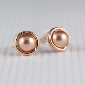 May include: A pair of rose gold stud earrings with a small, round, peach-coloured pearl in the centre of each earring.