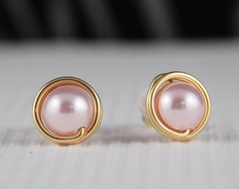 Può includere: Un paio di orecchini a lobo con perle rotonde rosa chiaro centrate all'interno di una cornice circolare in filo metallico color oro. Il design presenta una perla liscia e lucida racchiusa da un singolo anello di filo d'oro, creando una silhouette circolare semplice per ogni orecchino.