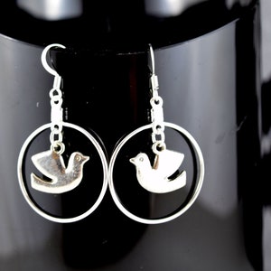 May include: Silver-toned earrings featuring a dove design within a circular frame. The earrings have a hook closure and are displayed against a black background with a decorative pattern.