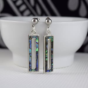 May include: A pair of silver earrings featuring a rectangular abalone shell design. The earrings have a silver post and a silver dangle with a rectangular frame. The abalone shell is iridescent with shades of blue, green, and purple.