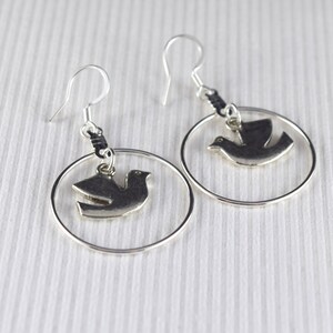 May include: A pair of silver-toned earrings featuring a dove pendant within a circular frame. The earrings have a hook closure and are displayed against a white background with subtle vertical lines. The dove is in flight.
