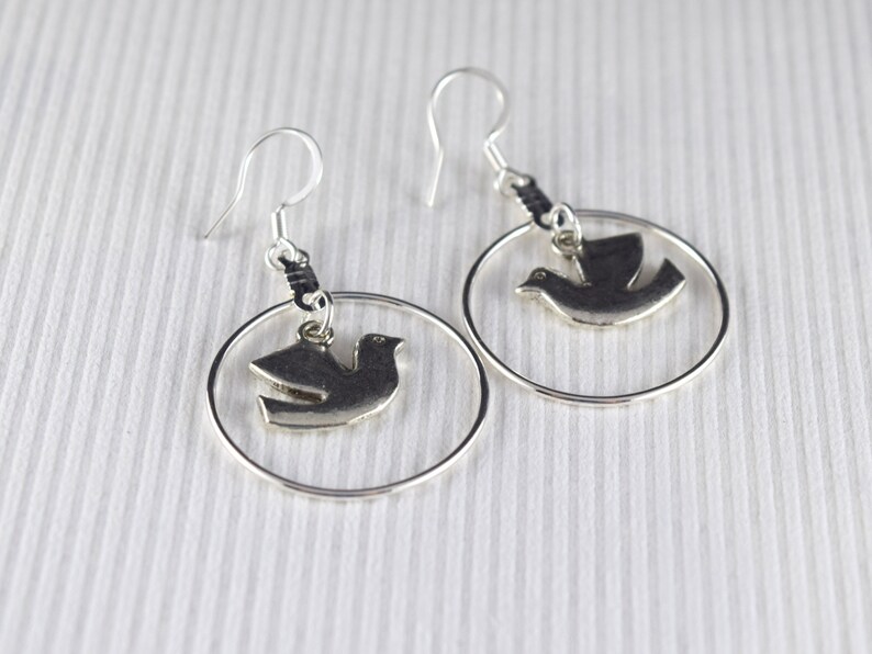 May include: A pair of silver-toned earrings featuring a dove pendant within a circular frame. The earrings have a hook closure and are displayed against a white background with subtle vertical lines. The dove is in flight.