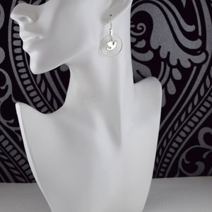 May include: Silver dove earrings. Each earring features a small, silver dove pendant suspended within a delicate silver hoop. The earrings are displayed on a white mannequin head against a black and white patterned background.