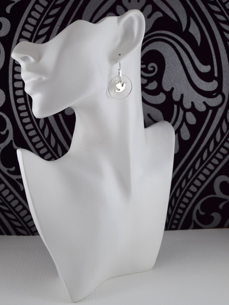 May include: Silver dove earrings. Each earring features a small, silver dove pendant suspended within a delicate silver hoop. The earrings are displayed on a white mannequin head against a black and white patterned background.