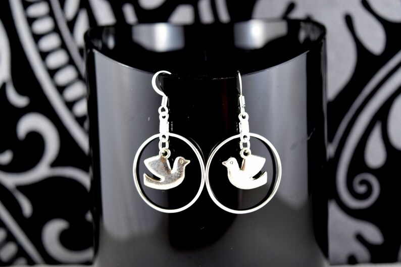 May include: Silver-toned earrings featuring a dove design within a circular frame. The earrings have a hook closure and are displayed against a black background with a decorative pattern.