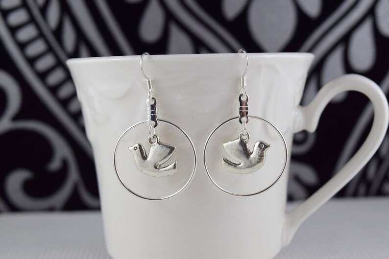 May include: A pair of silver-toned hoop earrings. Each earring features a small, detailed dove charm suspended within a circular frame. The earrings are displayed against a white mug, with a patterned background.