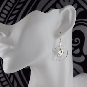May include: Silver-toned earrings featuring a dove pendant within a circular hoop. The earrings are displayed on a white mannequin head against a black and white patterned background. The dove pendant is a stylized representation of a bird.