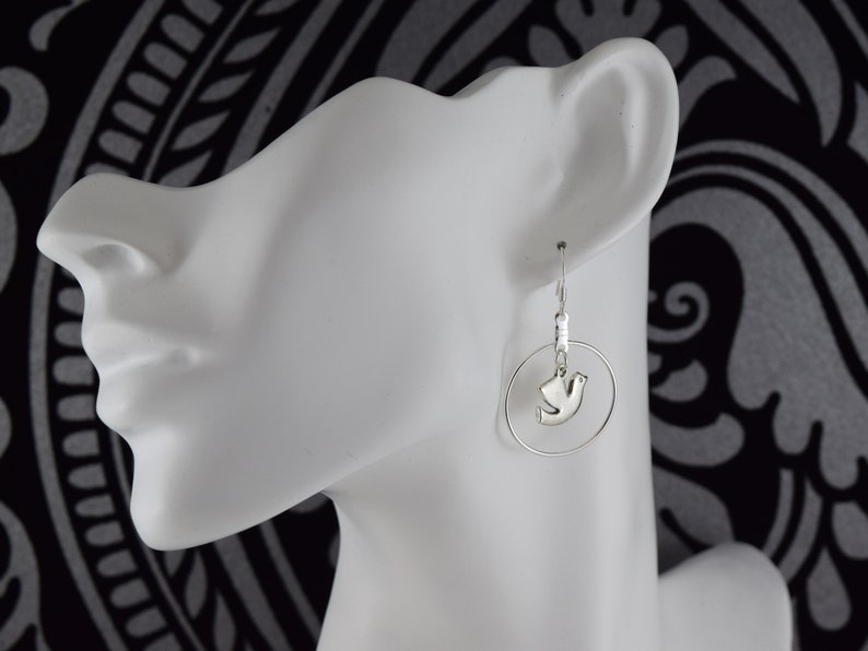 May include: Silver-toned earrings featuring a dove pendant within a circular hoop. The earrings are displayed on a white mannequin head against a black and white patterned background. The dove pendant is a stylized representation of a bird.