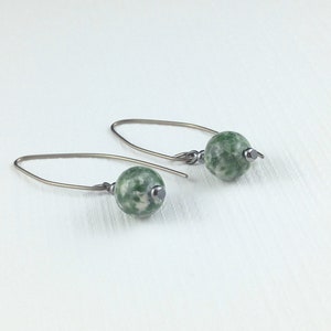 Peut inclure: Une paire de boucles d'oreilles pendantes composées de perles rondes en pierre marbrée verte et blanche, suspendues à des crochets métalliques foncés incurvés. Chaque boucle comporte une petite perle d'espacement métallique sous la pierre.