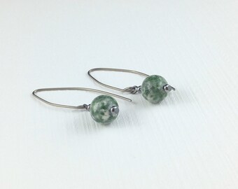 Può includere: Un paio di orecchini pendenti con perle rotonde in pietra screziata verde e bianca, sospese a ganci metallici scuri e ricurvi. Ogni orecchino presenta una piccola perlina distanziatrice in metallo scuro posizionata sotto la sfera di pietra.