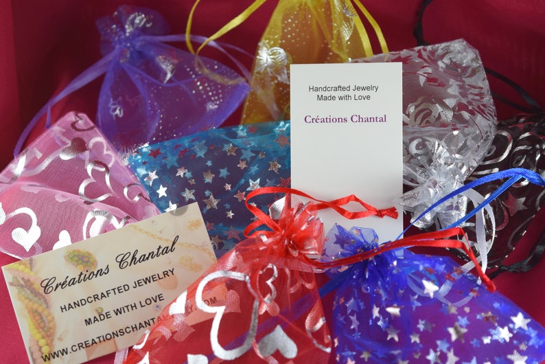 May include: An assortment of small, colorful organza bags filled with jewelry, some adorned with star and heart patterns. The image includes business cards with the text "Cr&eacute;ations Chantal, Handcrafted Jewelry, Made with Love."