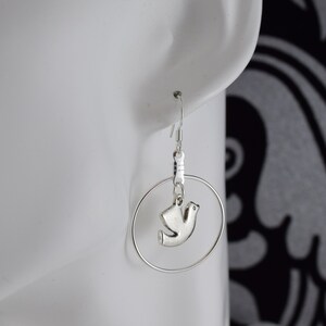 May include: Silver-toned hoop earrings with a small dove charm. The earrings feature a simple hook closure and a circular hoop design. The dove charm is a stylized representation of a bird, adding a touch of elegance to the jewelry.