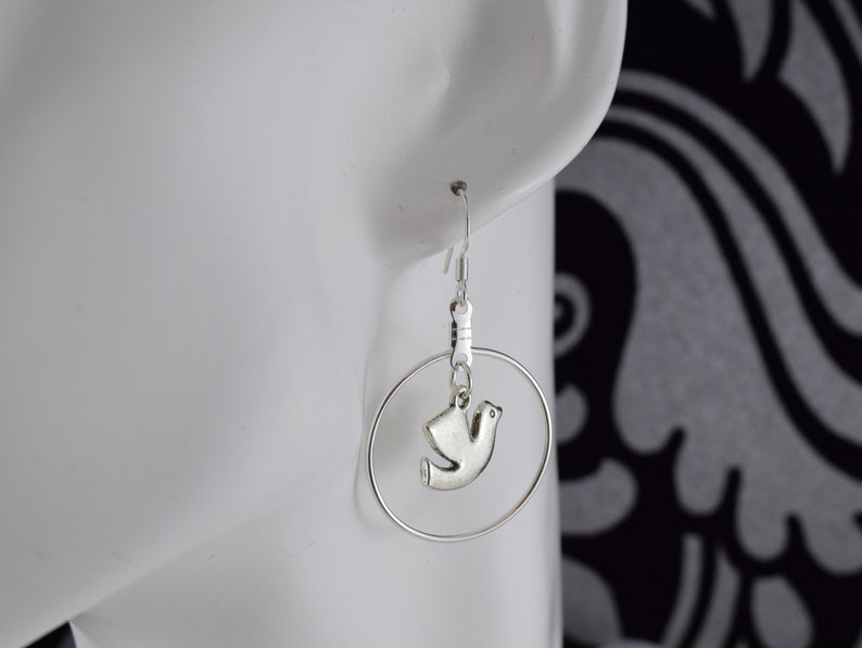 May include: Silver-toned hoop earrings with a small dove charm. The earrings feature a simple hook closure and a circular hoop design. The dove charm is a stylized representation of a bird, adding a touch of elegance to the jewelry.