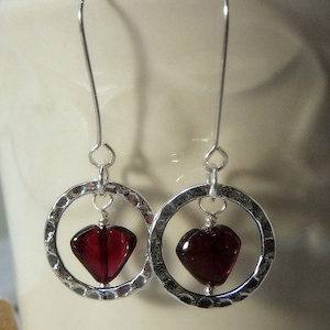 May include: A pair of silver-toned earrings. Each earring features a hammered silver hoop with a deep red, heart-shaped bead suspended in the center. The earrings have a simple hook design for pierced ears.