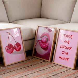 May include: Three framed wall art prints with light wood frames. The first print features two glossy red cherries on a pink background. The second shows bright pink lips holding a lollipop. The third has the red text TAKE ME DRUNK I'M HOME.