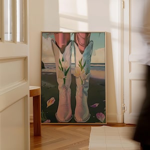 May include: A framed art print featuring a pair of white cowboy boots filled with white tulips, set against a beach scene with a light blue sky and ocean. The boots have a subtle pink hue, and the artwork is framed in a light wood.