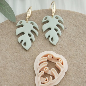 May include: A pair of light green Monstera leaf earrings with gold-colored hooks. A pink leaf-shaped cutter is also visible. The earrings and cutter are on a neutral-colored surface. The earrings are a tropical-themed accessory.