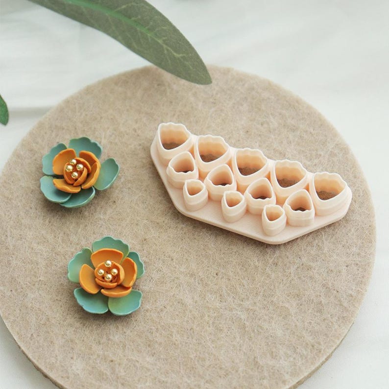 May include: Two handmade floral embellishments with teal and orange petals and a light pink plastic flower cutter on a tan felt surface. The flowers have small gold bead centres. The cutter has various flower shapes.