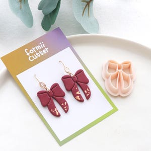 May include: Burgundy bow earrings with gold accents and a matching clay cutter. The earrings have a gold hook and a gold leaf design. The card behind the earrings has the text "formii Cutter".