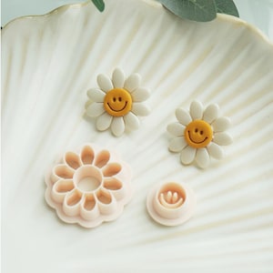 May include: A collection of daisy-shaped cookie cutters and decorative accents. The cutters are light pink, with one resembling a daisy and another with a smiley face. Two daisy accents feature white petals and yellow smiley faces. The items are arranged on a white, scalloped surface.