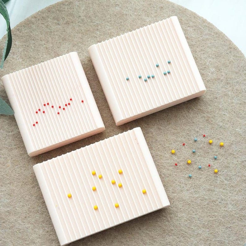 May include: Three light pink, rectangular, ridged soap dishes with colorful dots. One dish has red dots, another has blue-green dots, and the third has yellow dots. A few loose dots are scattered nearby.