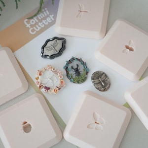 Può includere: Una collezione di fustelle artigianali e stampi decorativi a tema insetti. Le fustelle sono quadrate e rosa chiaro, con disegni di libellule e farfalle. Gli stampi presentano dettagliati disegni di insetti e floreali. Il testo "Formii Cutter" è visibile.