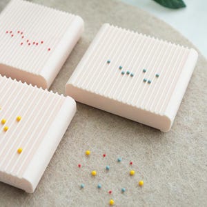 May include: Three light pink, rectangular soap dishes with vertical ridges. Each dish has a pattern of small, colorful beads on top. Some beads are red, yellow, and blue. The dishes are on a light gray surface.