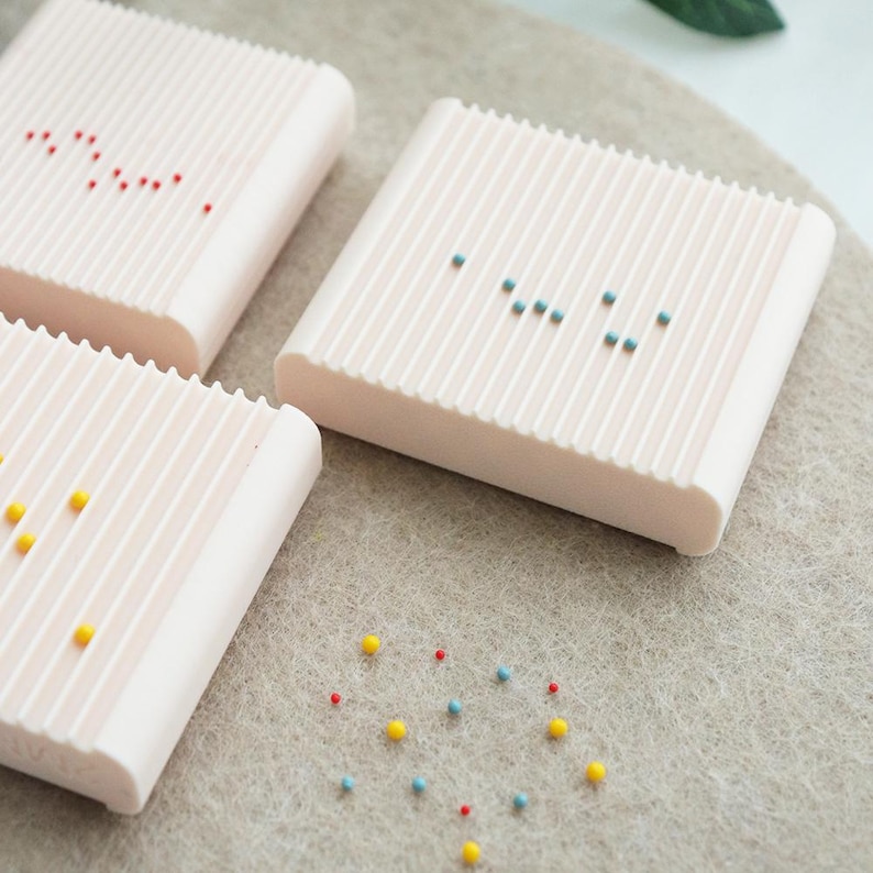 May include: Three light pink, rectangular soap dishes with vertical ridges. Each dish has a pattern of small, colorful beads on top. Some beads are red, yellow, and blue. The dishes are on a light gray surface.