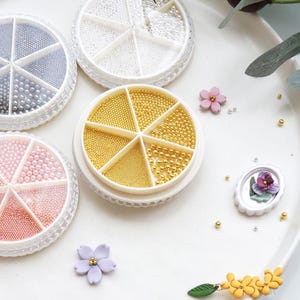 Puede incluir: Pequeñas cuentas redondas de varios colores, incluyendo oro, plata y rosa, se muestran en recipientes circulares divididos. Acentos florales decorativos y un diseño floral en miniatura enmarcado sugieren suministros de artesanía.