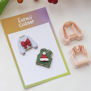 Könnte beinhalten: Ein Bild mit weihnachtlichen Ausstechformen und dekorierten Keksen. Die Kekse sind wie Pullover geformt, einer hellblau mit roter Schleife und der andere grün mit Weihnachtsmütze. Die Ausstecher sind aus rosa Kunststoff und haben Pulloverform.