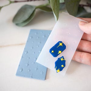 May include: A light blue silicone mold with a star pattern and two blue and yellow star-shaped clay earrings. The earrings are in a clear plastic mold. The background is white.