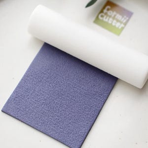 Op de afbeelding: Een witte deegroller ligt op een getextureerd, vierkant, periwinkleurig oppervlak. De afbeelding toont ook een kleine, rechthoekige sticker met de woorden "Formii Cutter" in handschrift.