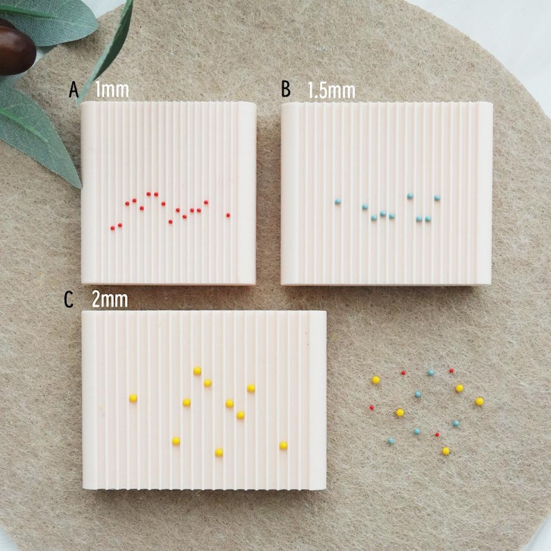 May include: Three small, textured, off-white rectangular containers with vertical lines, each holding small, colorful beads. The containers are labeled with the text "A 1mm", "B 1.5mm", and "C 2mm". A scattering of beads is also present.