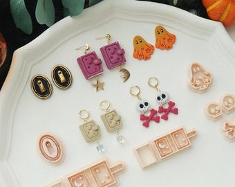 May include: A collection of Halloween-themed earrings and craft supplies. Includes earrings shaped like ghosts, skulls, books, and oval shapes. The color palette features pink, orange, gold, and black. Also includes craft molds for making Halloween-themed items.