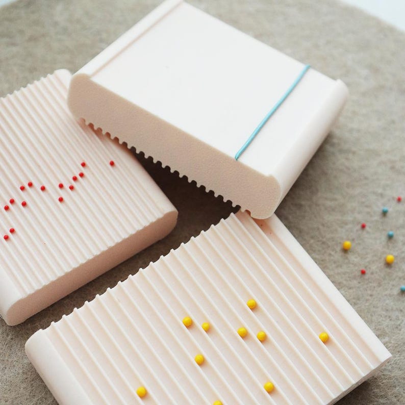 May include: Three light pink soap dishes with a ridged design. One dish has a blue elastic band, and the other two have small red and yellow decorative beads. The dishes are rectangular with rounded edges and a textured surface.