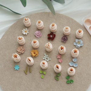 May include: A collection of light pink flower-shaped fondant cutters and various colorful sugar flowers and leaves. The cutters are arranged on a beige surface, alongside the decorative floral elements, perfect for cake decorating.