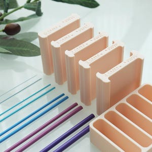 May include: A collection of peach-colored knitting tools, including rectangular gauge swatches labeled with measurements in millimeters, and a variety of colorful knitting needles. The tools are arranged on a light-colored surface.