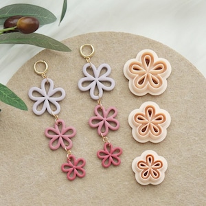 Op de afbeelding: Sieraden en knutselbenodigdheden met bloementhema. Twee paar bloemvormige oorbellen in lavendel- en roze tinten, met goudkleurige ringen en connectoren. Ook drie bloemvormige koekjessnijders.