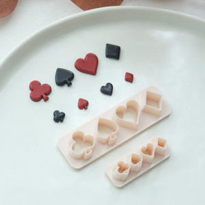 Puede incluir: Formas de naipes y cortadores de galletas a juego. Los palos incluyen corazones, diamantes, tréboles y picas en rojo y negro. Los cortadores de galletas son de color rosa claro.