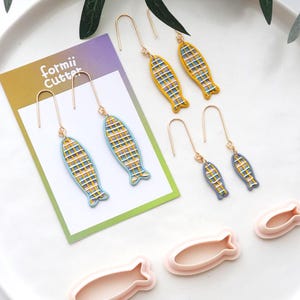 Puede incluir: Pendientes con diseño de pez en amarillo, azul y blanco, con ganchos dorados. Los pendientes se muestran sobre una superficie blanca con una tarjeta que dice "formii Cutter". También se incluyen cortadores rosas con forma de pez.