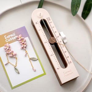 May include: A light pink and brown Formii Cutter tool with a chain and a small brown circle. Next to it are pink flower earrings with gold chains and green leaf accents. The earrings are on a white card with the text "Formii Cutter".
