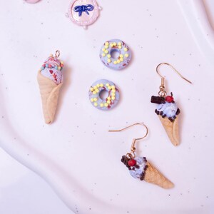 May include: A collection of miniature food-themed jewelry and charms. The image features two ice cream cone earrings, several miniature donuts, and a pink bow-shaped charm. The earrings and donuts have colorful sprinkles.