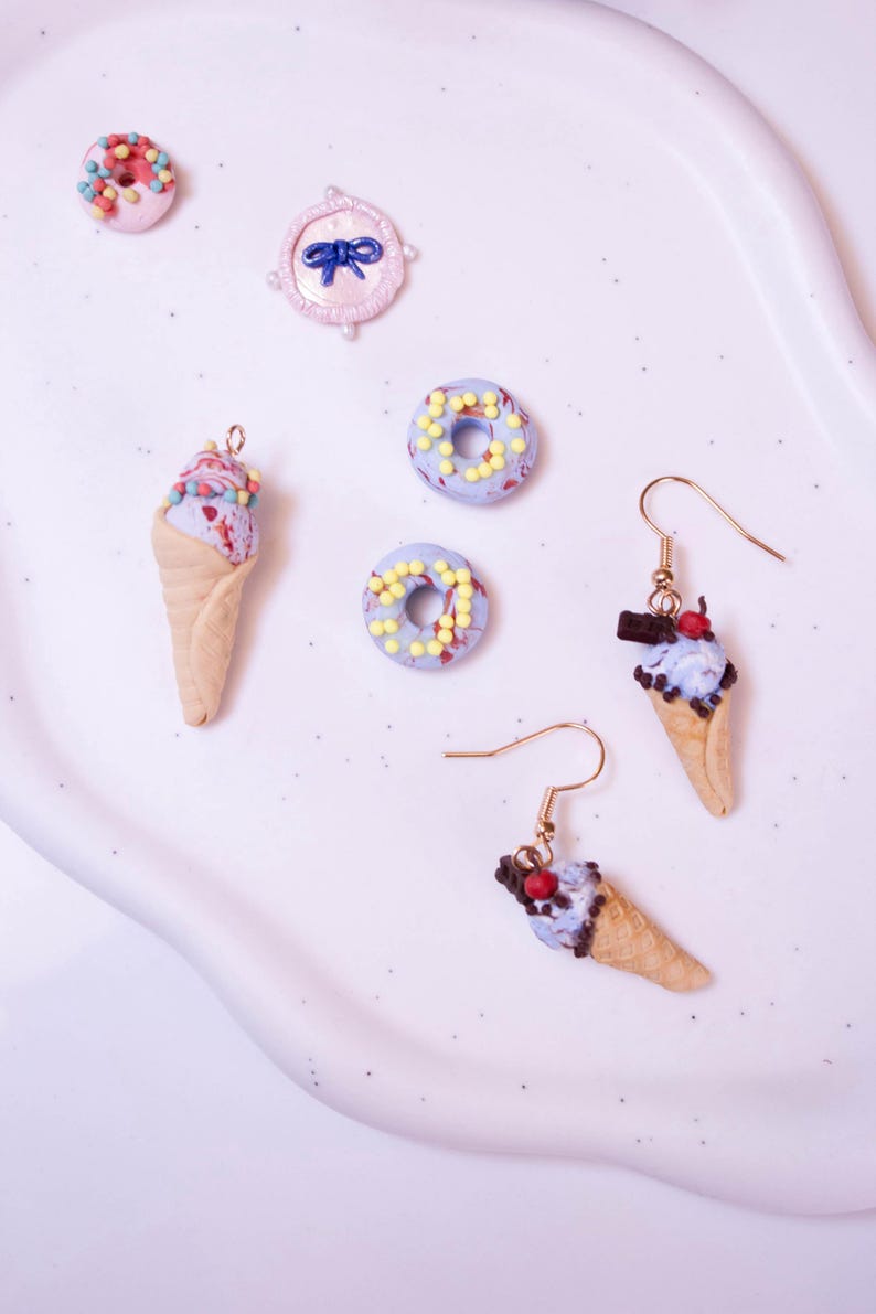 May include: A collection of miniature food-themed jewelry and charms. The image features two ice cream cone earrings, several miniature donuts, and a pink bow-shaped charm. The earrings and donuts have colorful sprinkles.