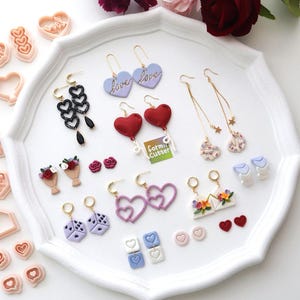 Op de afbeelding: Een verzameling handgemaakte oorbellen en kleisnijders gerangschikt op een wit dienblad. De oorbellen hebben hart-, dobbelsteen- en bloemontwerpen in verschillende kleuren. Sommige oorbellen hebben het woord "love" erop.