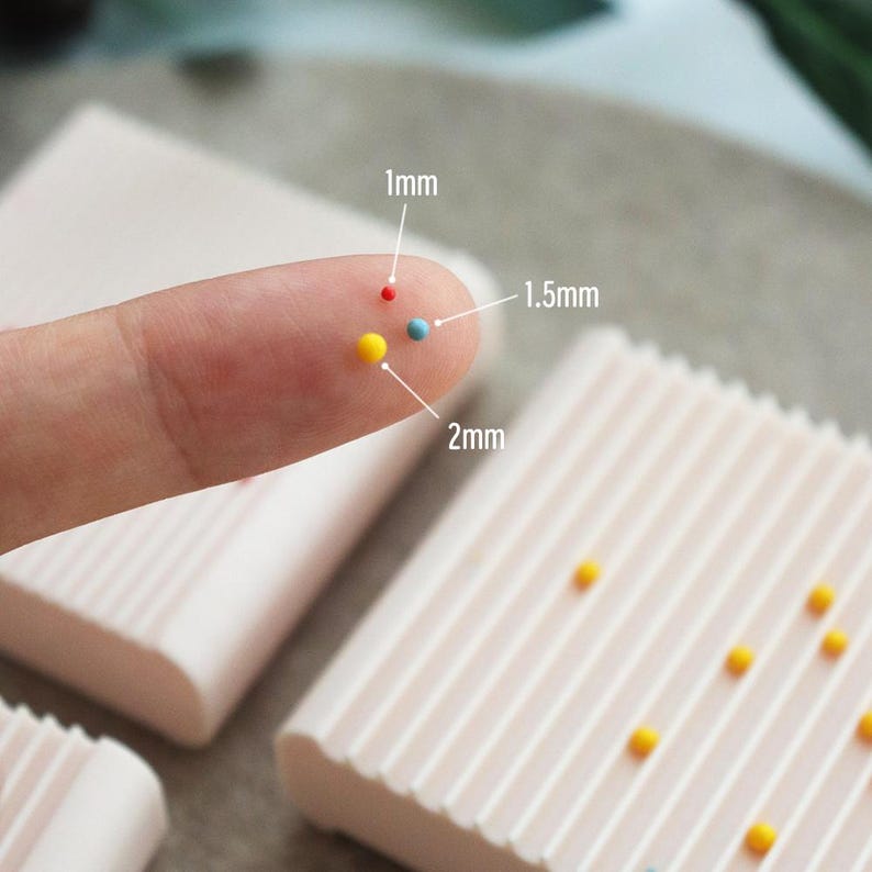 May include: Close-up of a finger holding small, colorful sprinkles. The sprinkles are red, blue, and yellow, with sizes labeled as 1mm, 1.5mm, and 2mm. Light pink, rectangular blocks with a ridged design are in the background.